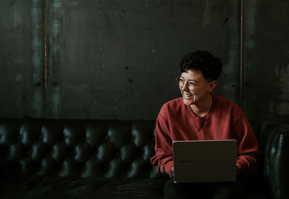 Smiling person with short, dark hair and glasses using laptop on a black leather sofa