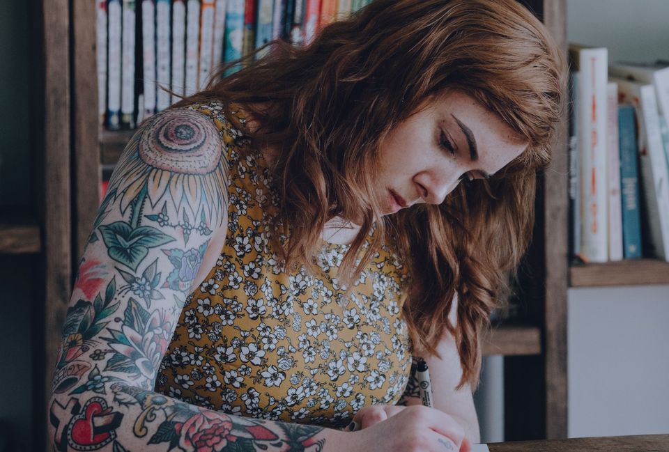 Long-haired woman with a yellow floral dress and colorful arm tattoos writing at a desk